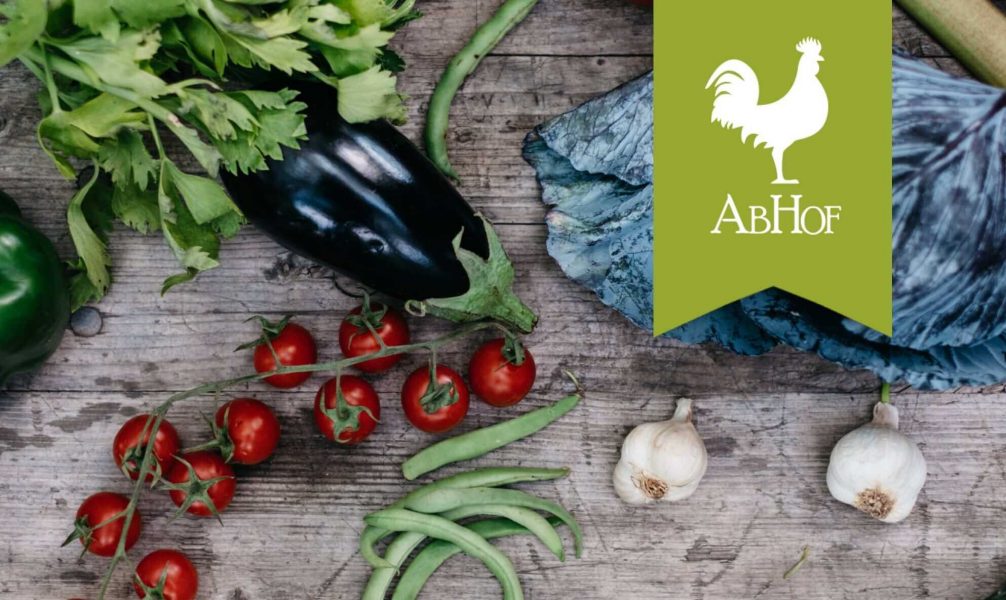 Die Marke Abhof die von einem weißen Hahn auf grünen Hintergrund dargestellt wird mit Gemüse auf einem Holztisch im Hintergrund